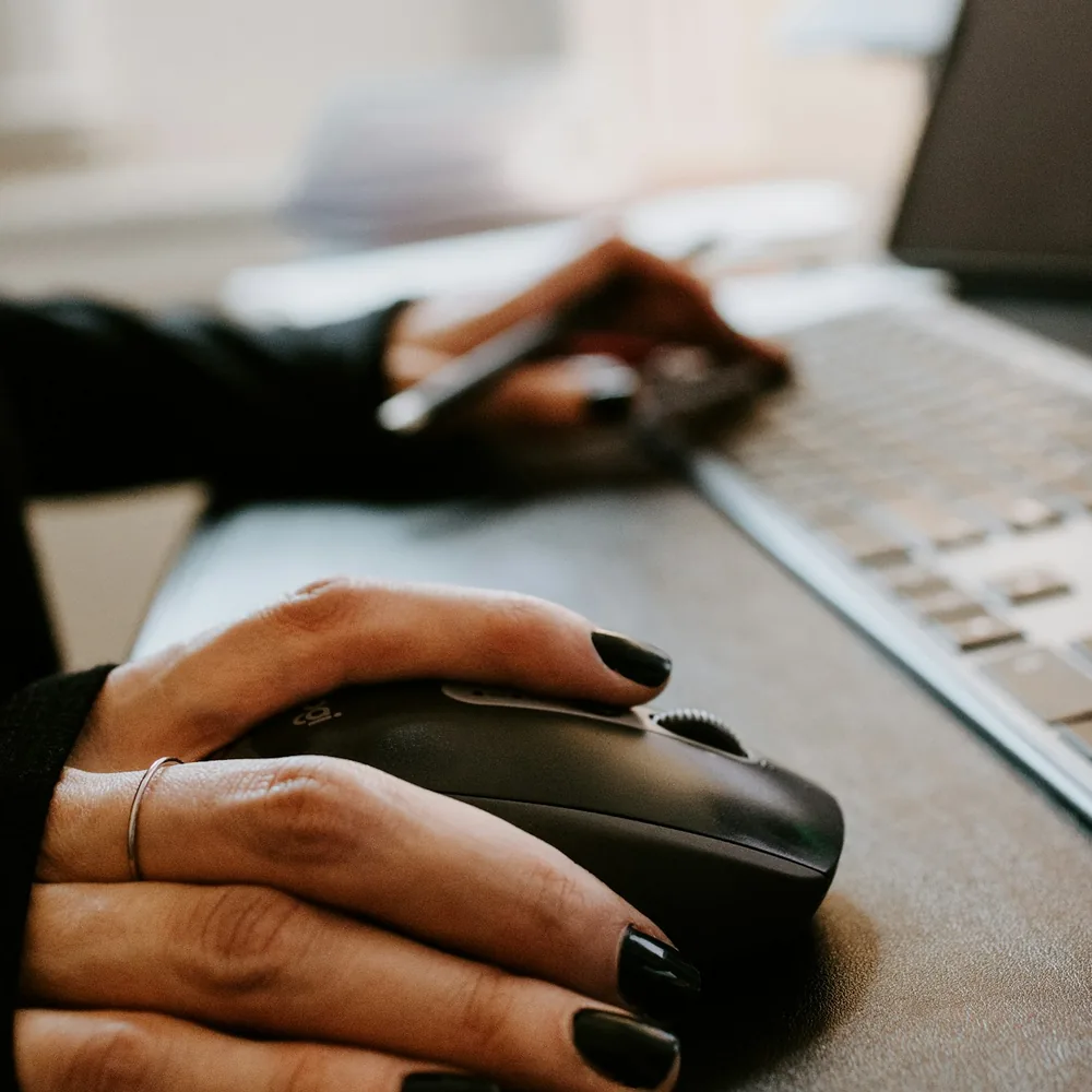 Hand on a black Logitech mouse on a desk beside a laptop keyboard, with a second hand holding a pen