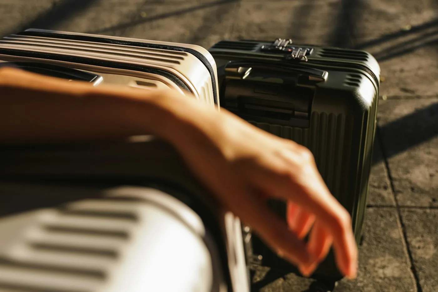 A hand reaching over two hardside suitcases, one white and one dark green, on a paved floor