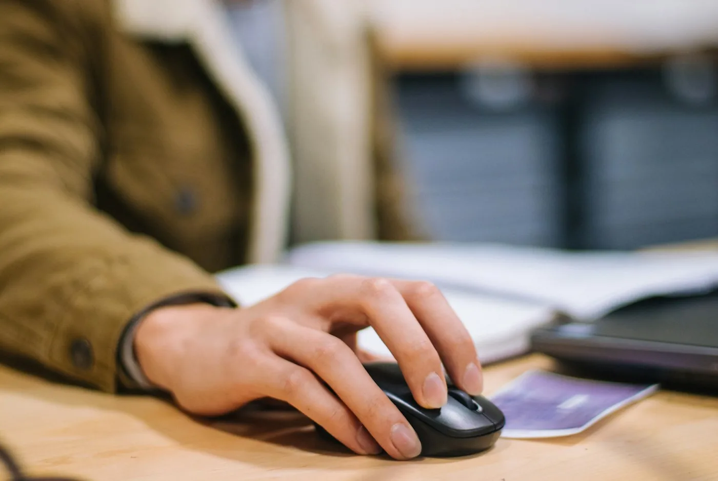 A hand resting on a black wireless mouse on a wooden desk beside a laptop and a notebook