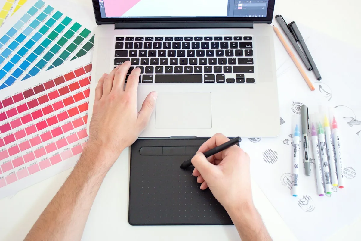 Hands drawing on a graphics tablet next to a laptop and color swatches, a design workspace