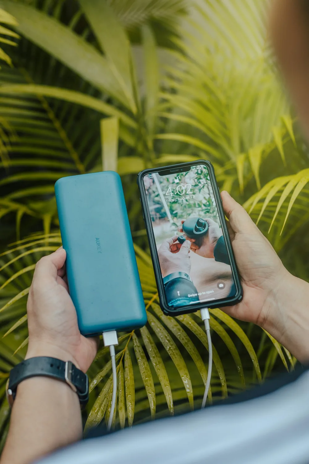 Zwei Hände halten eine grüne Powerbank und ein Smartphone vor grünen Palmblättern
