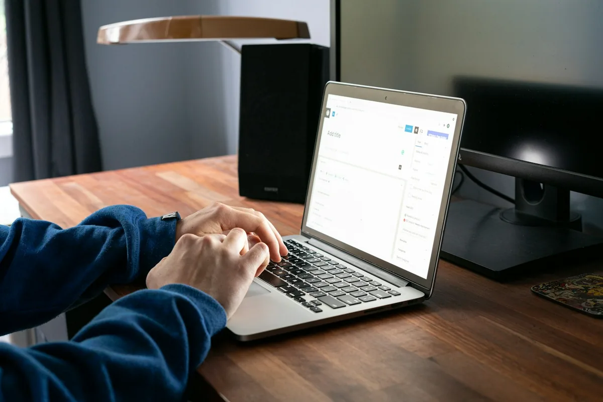 Hands typing on a laptop at a wooden desk under a lamp
