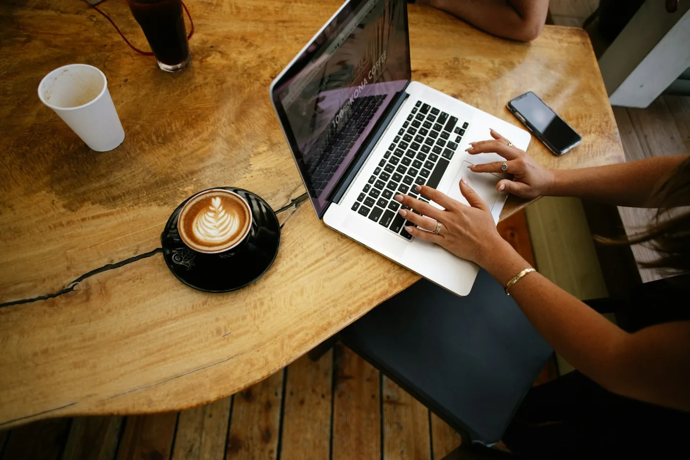 Hands typing on a laptop on a wooden table next to a latte and a phone