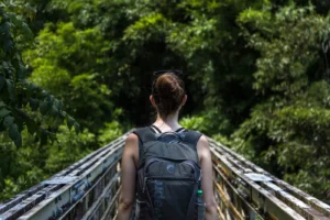 Hiker seen from behind with a backpack walking across a steel bridge in a green forest