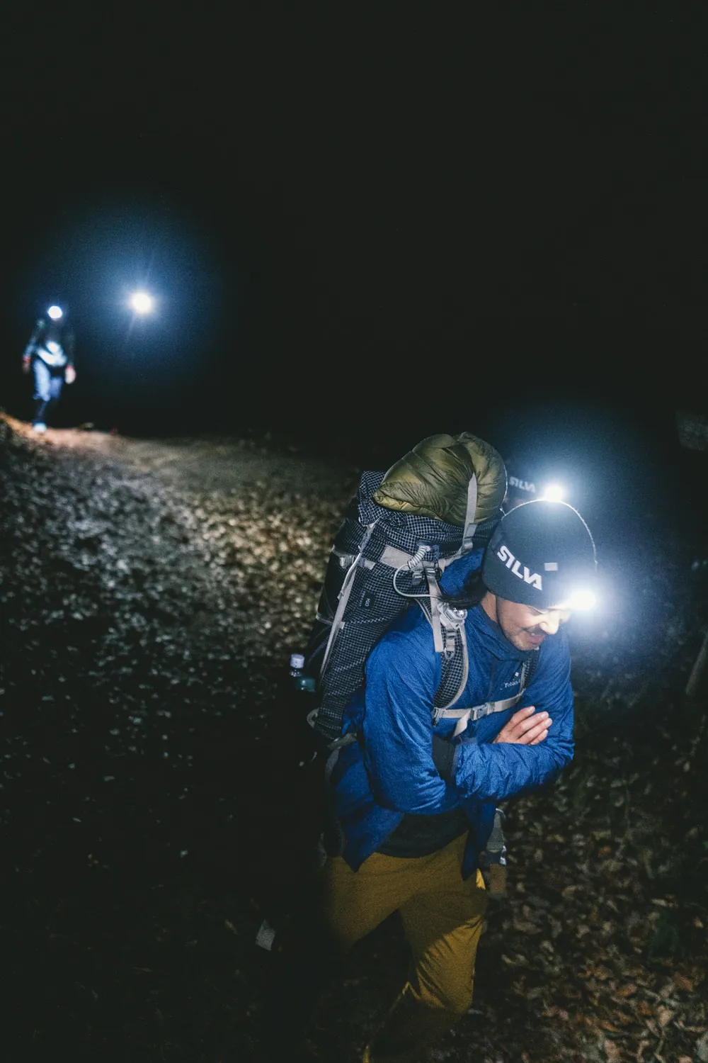Zwei Wanderer mit Stirnlampen auf einem dunklen Pfad, einer von ihnen trägt einen Rucksack
