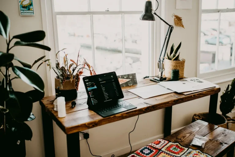 Wooden desk with a laptop, a lamp and plants next to a bright window