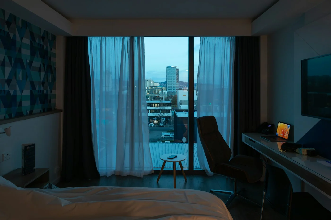Hotel room with a desk by a window, a laptop on it and a city view through sheer curtains
