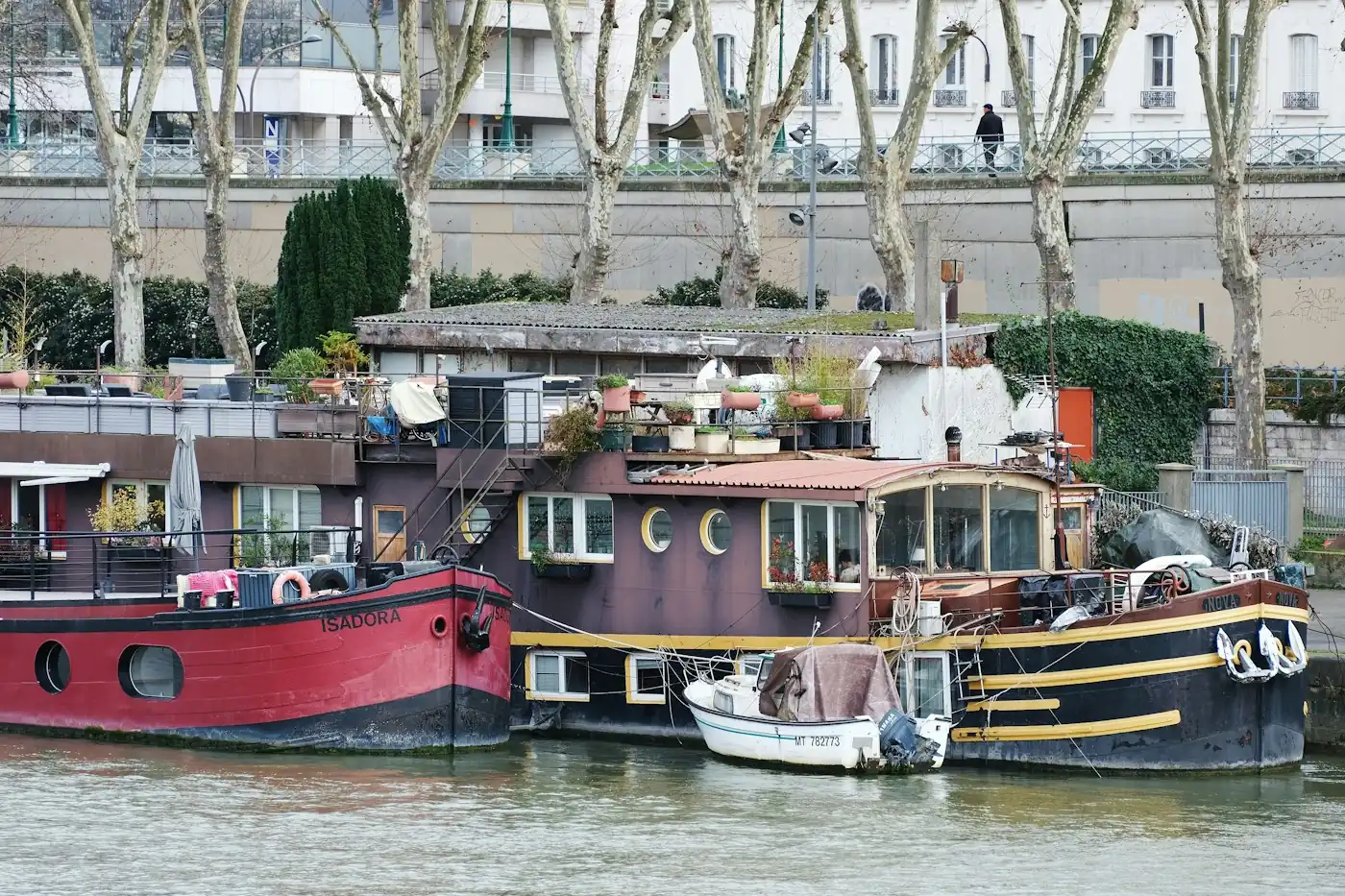 Houseboats moored side by side on a river in front of a tree-lined embankment
