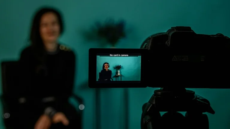 A camera monitor showing a woman being interviewed, with the woman blurred behind it against a teal wall