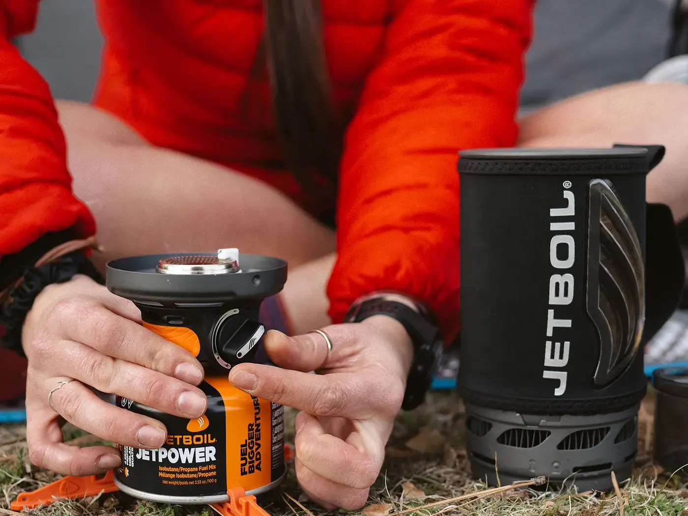 Hände befestigen einen Jetboil-Brenner an einer orangefarbenen JetPower-Gaskartusche auf dem Boden an einem Campingplatz
