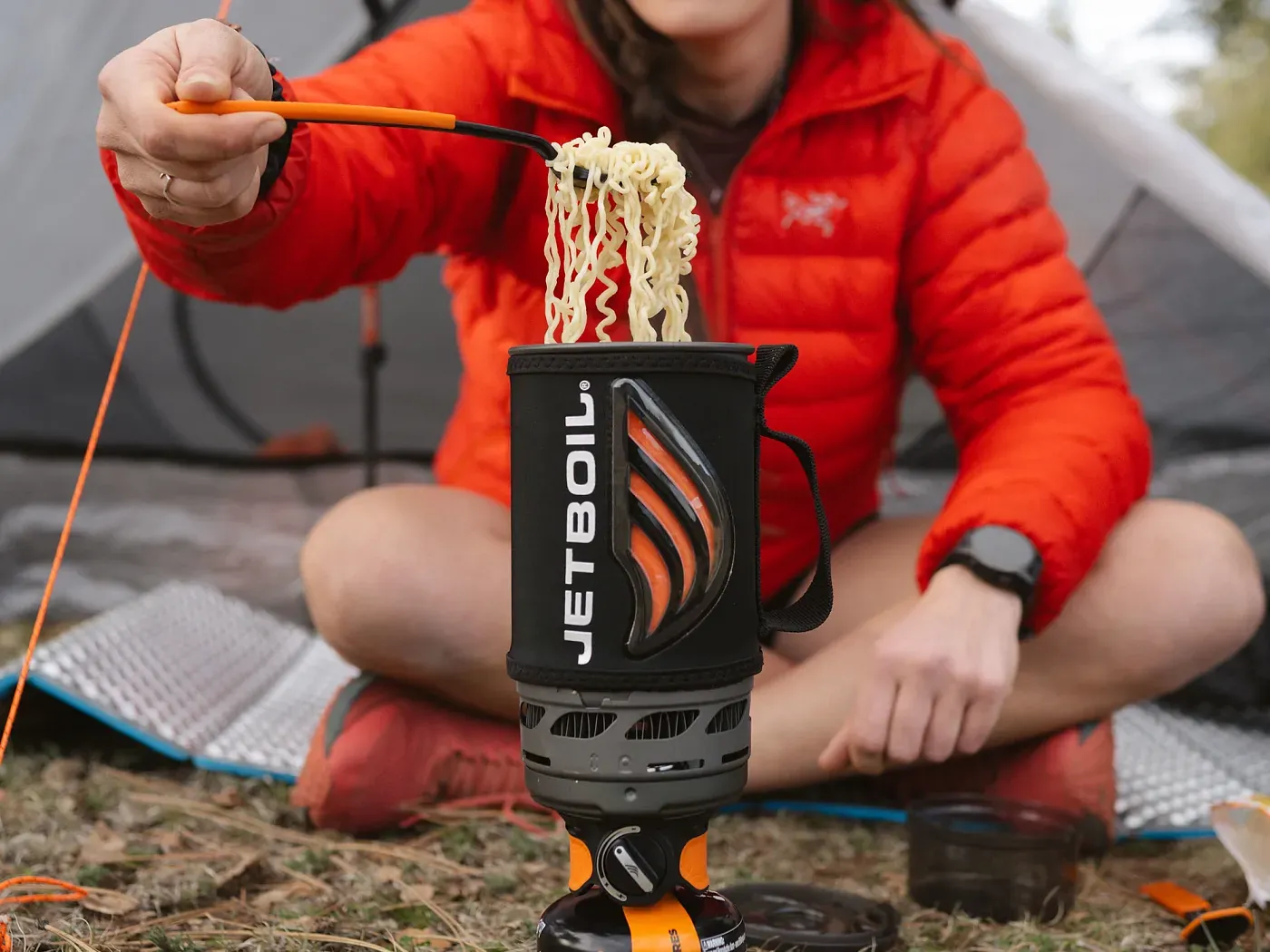 Lächelnder Camper in orangefarbener Jacke isst Nudeln aus einem Jetboil Flash-Kochtopf an einem Campingplatz