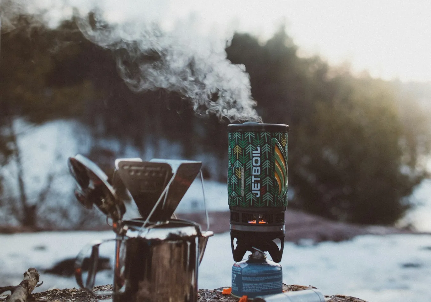 Dampf steigt von einem Jetboil-Kocher auf einem Felsen auf, neben einem Metallbecher in einem verschneiten Wald