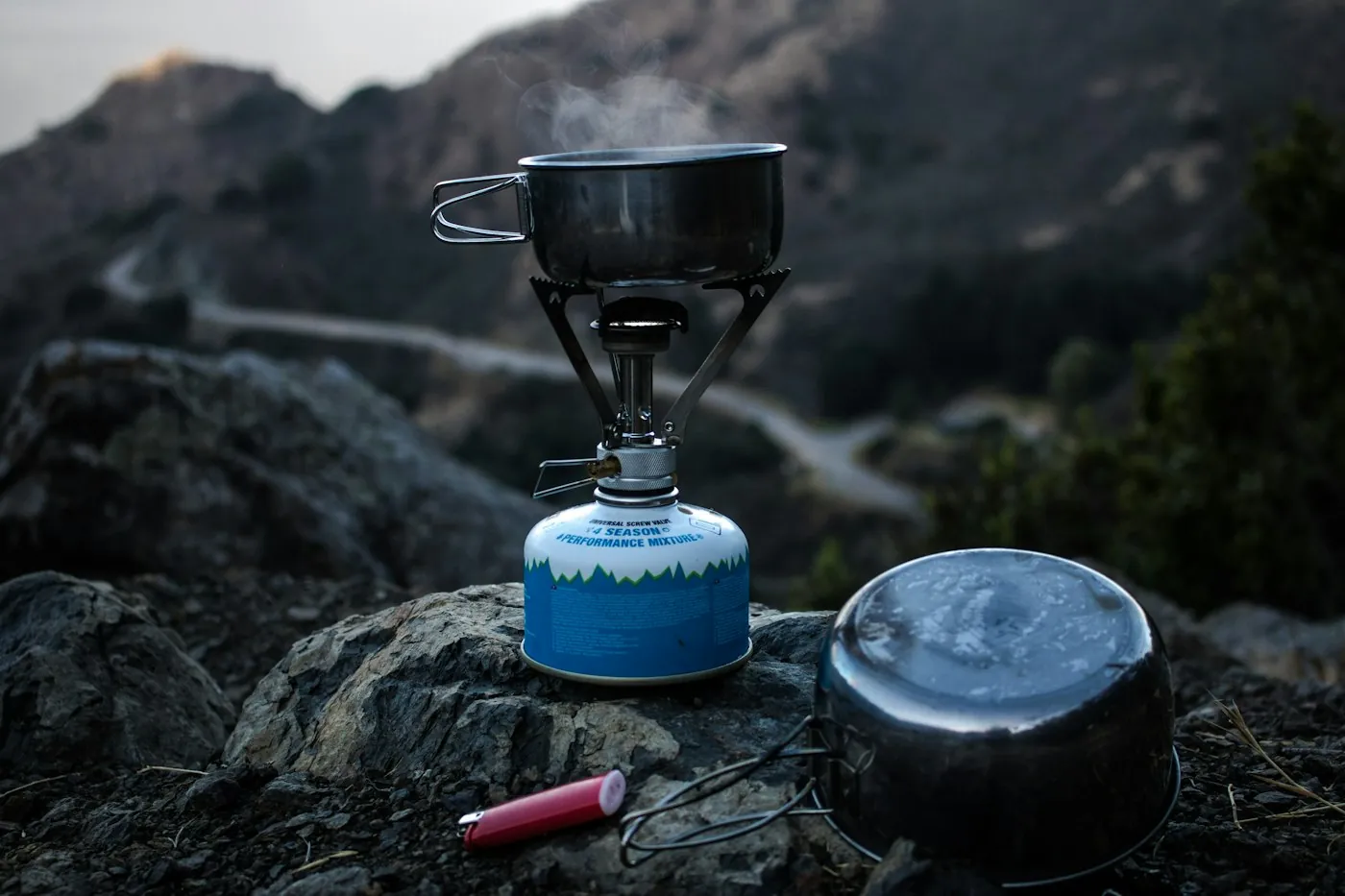 Small blue gas canister stove with a cooking pot on top, set on rocks above a winding mountain road