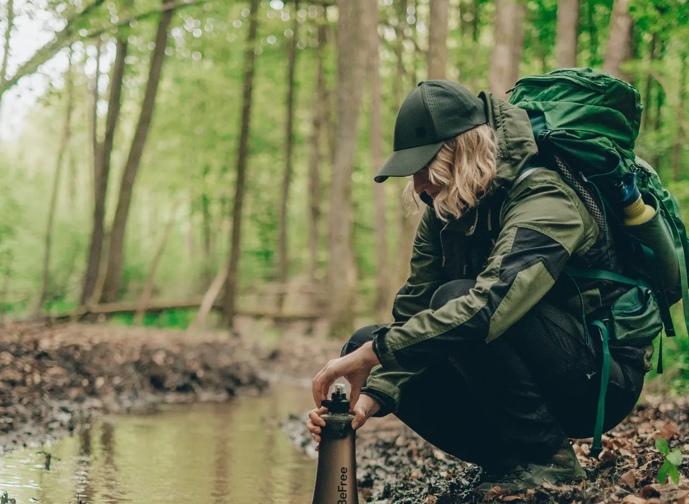 Wanderer in grüner Jacke und Kappe hockt an einem Waldbach und füllt eine dunkle Katadyn BeFree Flasche