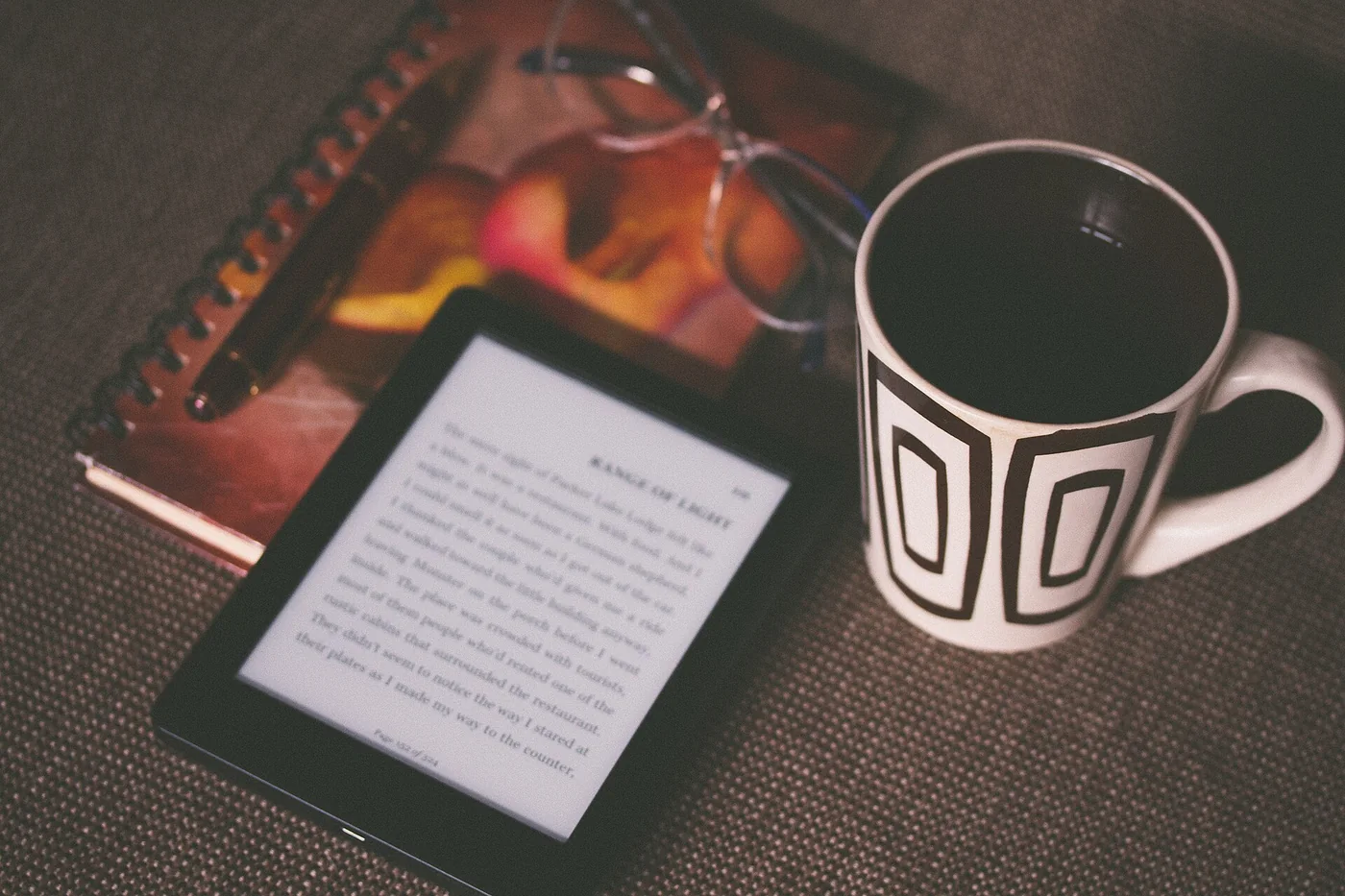 A Kindle e-reader lying on a notebook next to a black and white mug and a pair of glasses