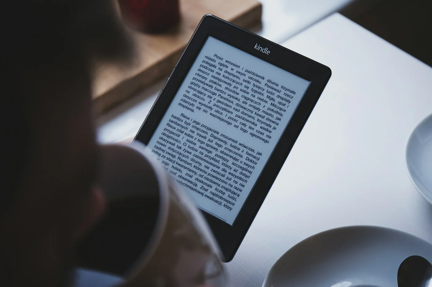 A Kindle e-reader tilted beside a coffee cup and saucer on a white table