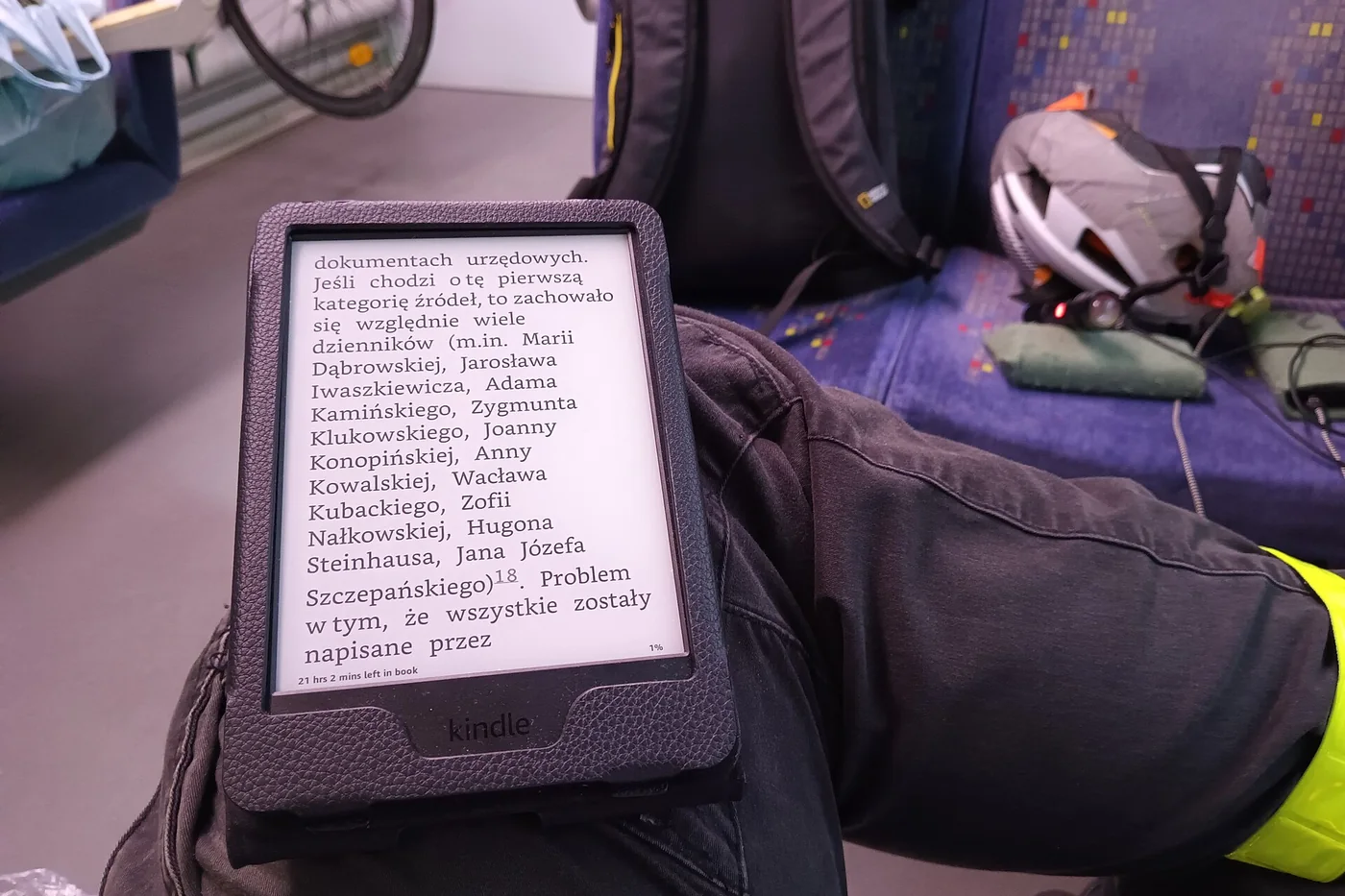 A Kindle in a black case held on a knee in a train compartment with a backpack and a helmet on the seat