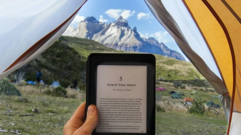 A hand holding a Kindle e-reader inside a tent that opens onto a mountain view