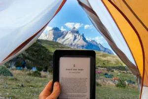 A hand holding a Kindle e-reader inside an open tent with snow-capped mountains outside
