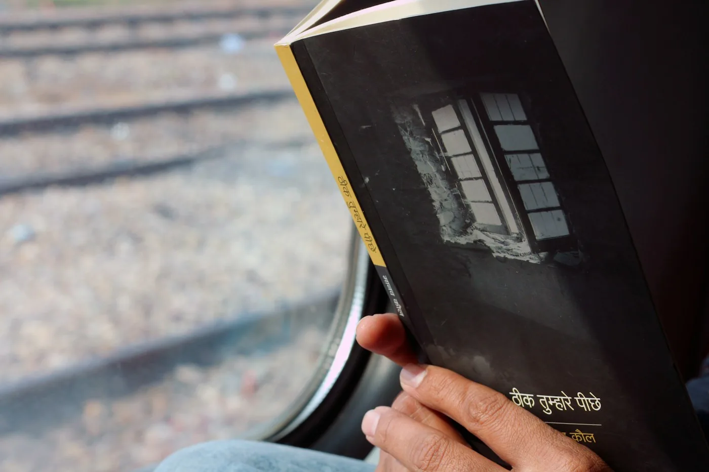 Una mano sosteniendo un libro abierto junto a la ventana de un tren, con vías de ferrocarril abajo
