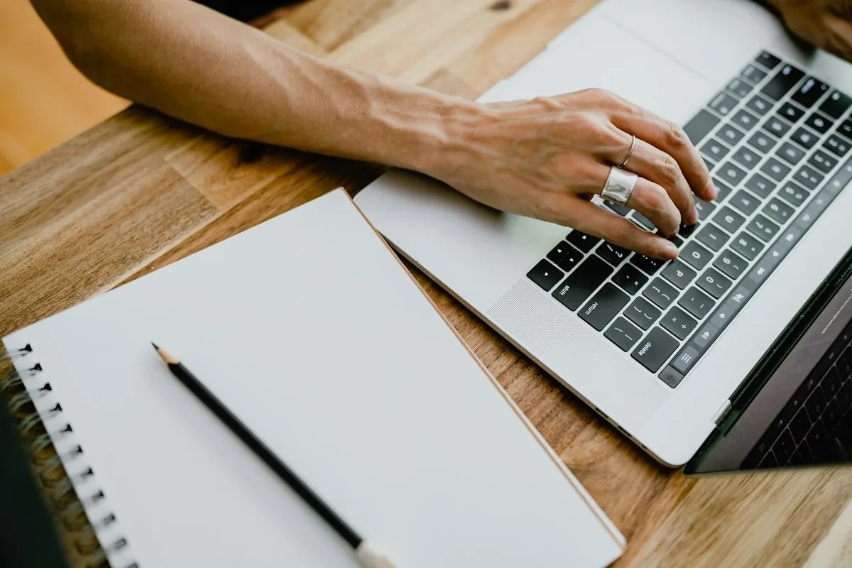 Hand on a laptop keyboard beside a blank notebook and pencil on a wooden table, planning the day with AI tools