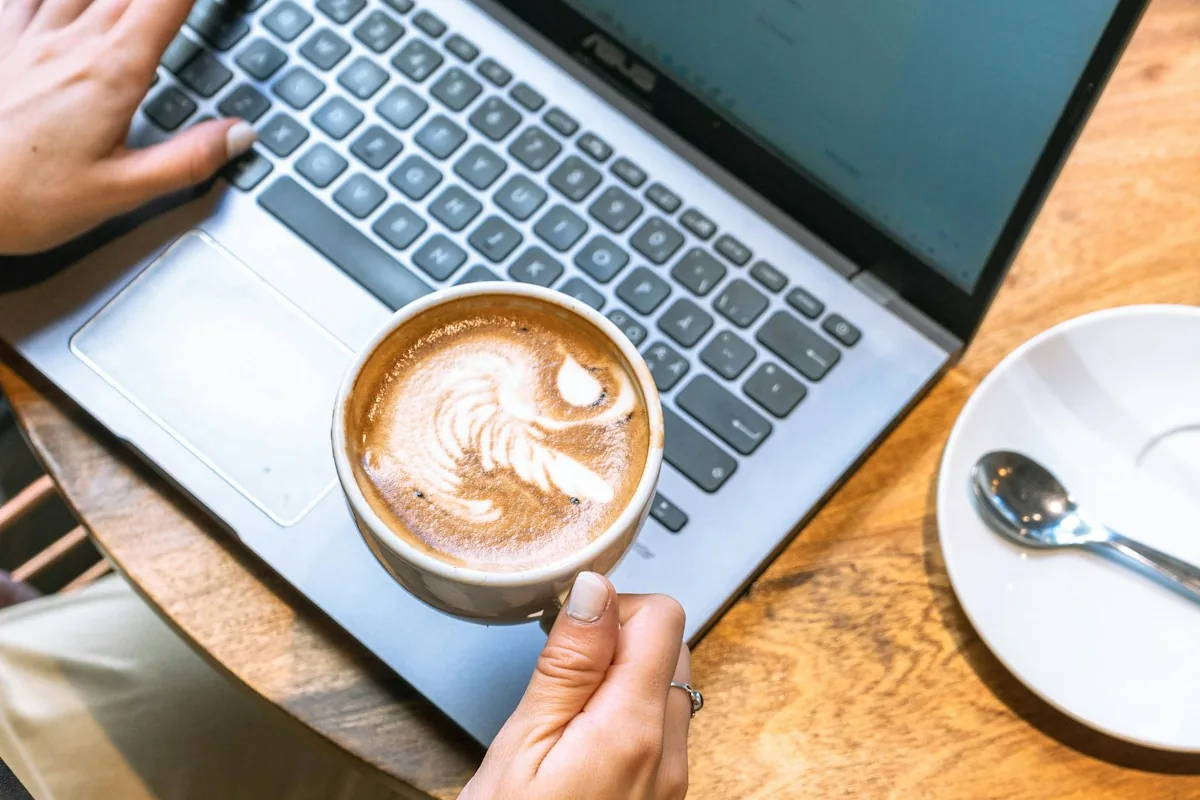 Hands on a laptop keyboard next to a latte on a wooden cafe table