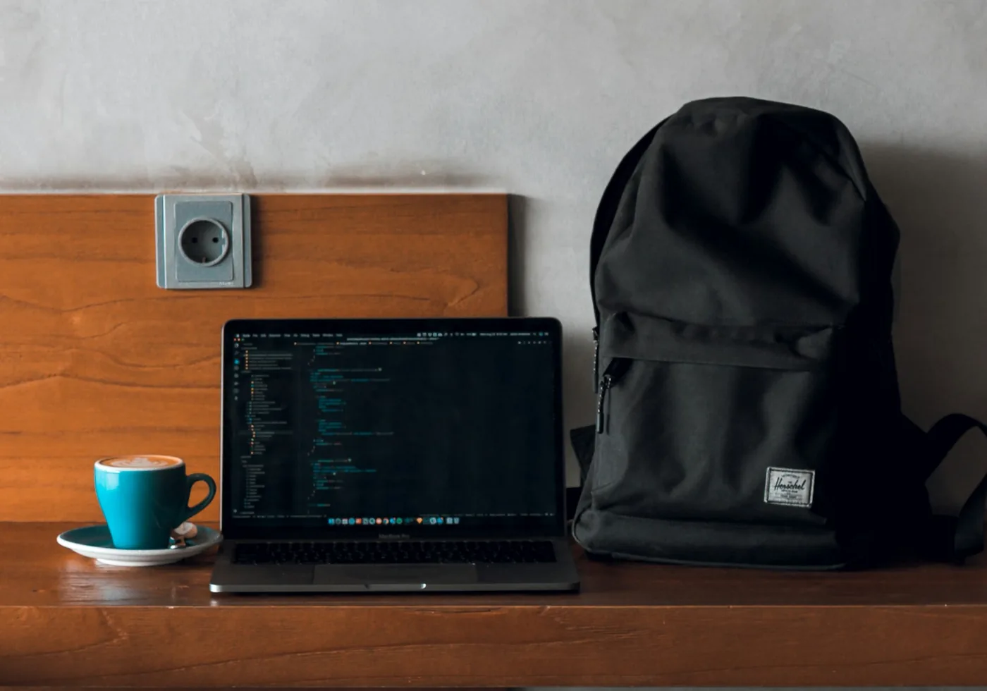 Geöffneter Laptop auf einem Holzregal neben einem schwarzen Rucksack und einer türkisen Kaffeetasse