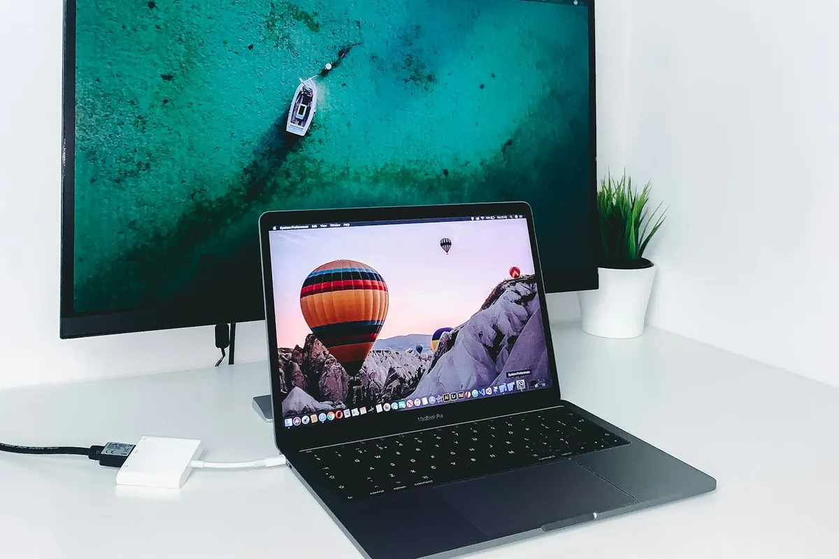 MacBook on a white desk with a USB-C adapter cable running to a larger monitor behind it
