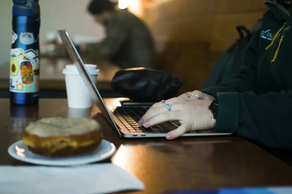 Person typing on a laptop at a cafe table with a coffee cup and a donut