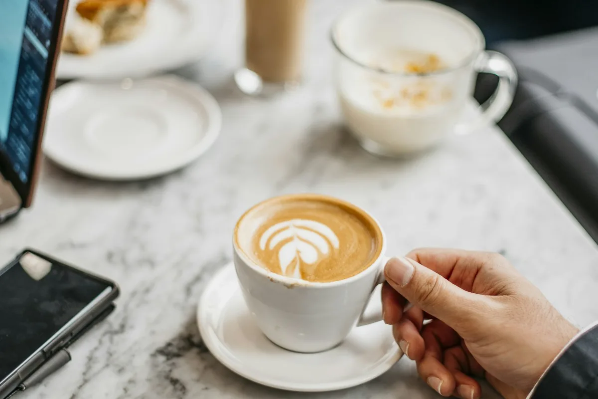 Een hand met een kopje latte op een marmeren cafétafel, met de rand van een laptop in de hoek