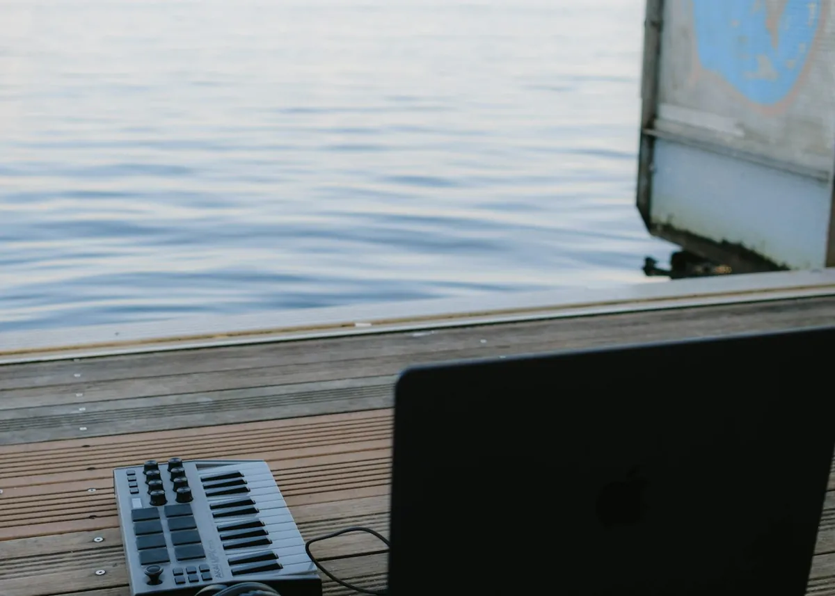 Laptop and a music keyboard on a wooden deck beside calm water