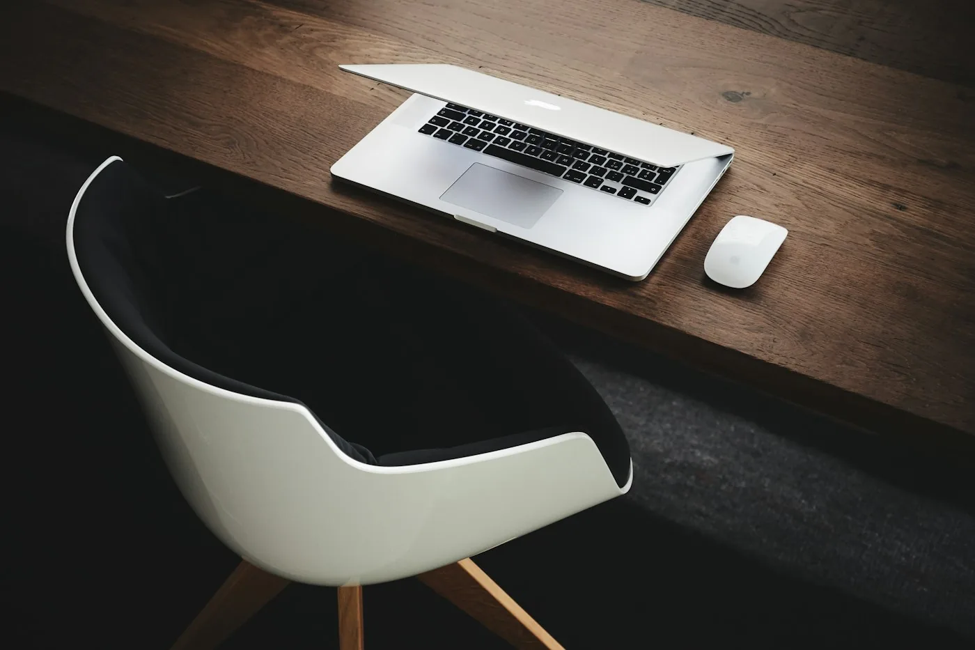 Open laptop and a white wireless mouse on a wooden desk next to a white chair