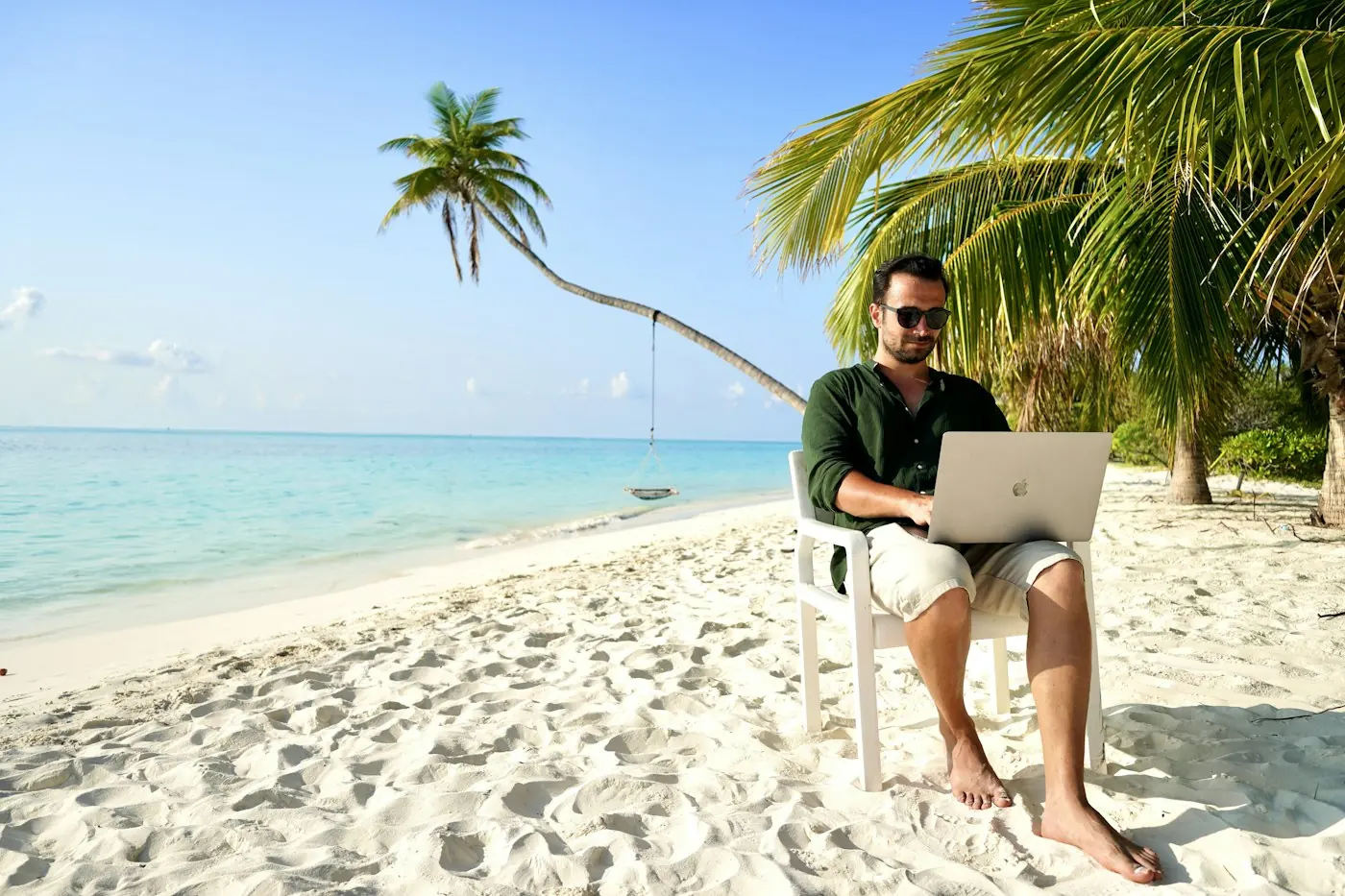 Man working on a laptop in a chair on a white sand beach with palm trees and turquoise water