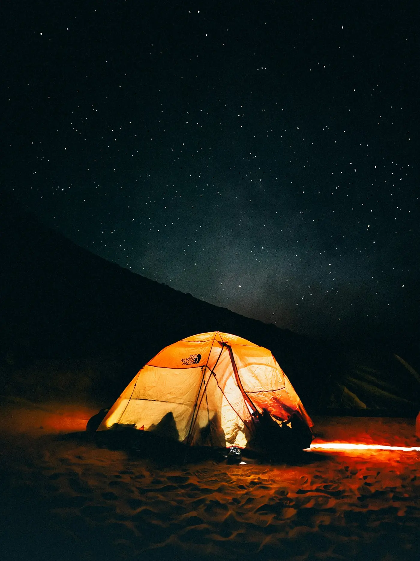 Ein orangefarbenes, von innen leuchtendes Zelt in einer dunklen Wüstenlandschaft unter einem Sternenhimmel
