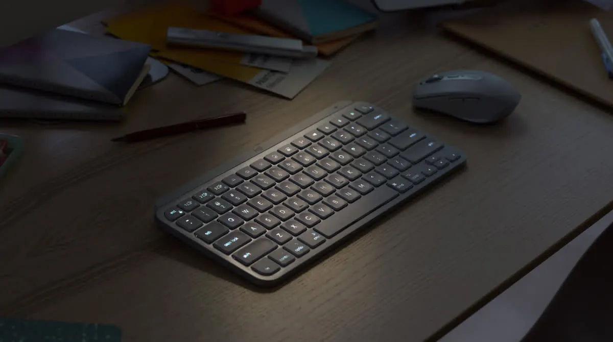 Logitech MX Keys Mini backlit keyboard on a dark wooden desk next to a mouse and notebooks