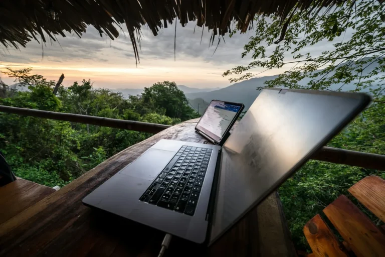 Open laptop on a wooden table on a hut deck with green hills and a sunset sky behind it