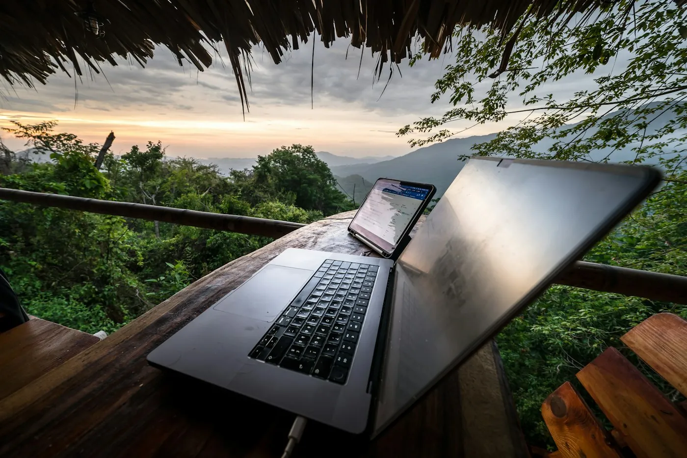 Open laptop on a wooden table on a hut deck with green hills and a sunset sky behind it
