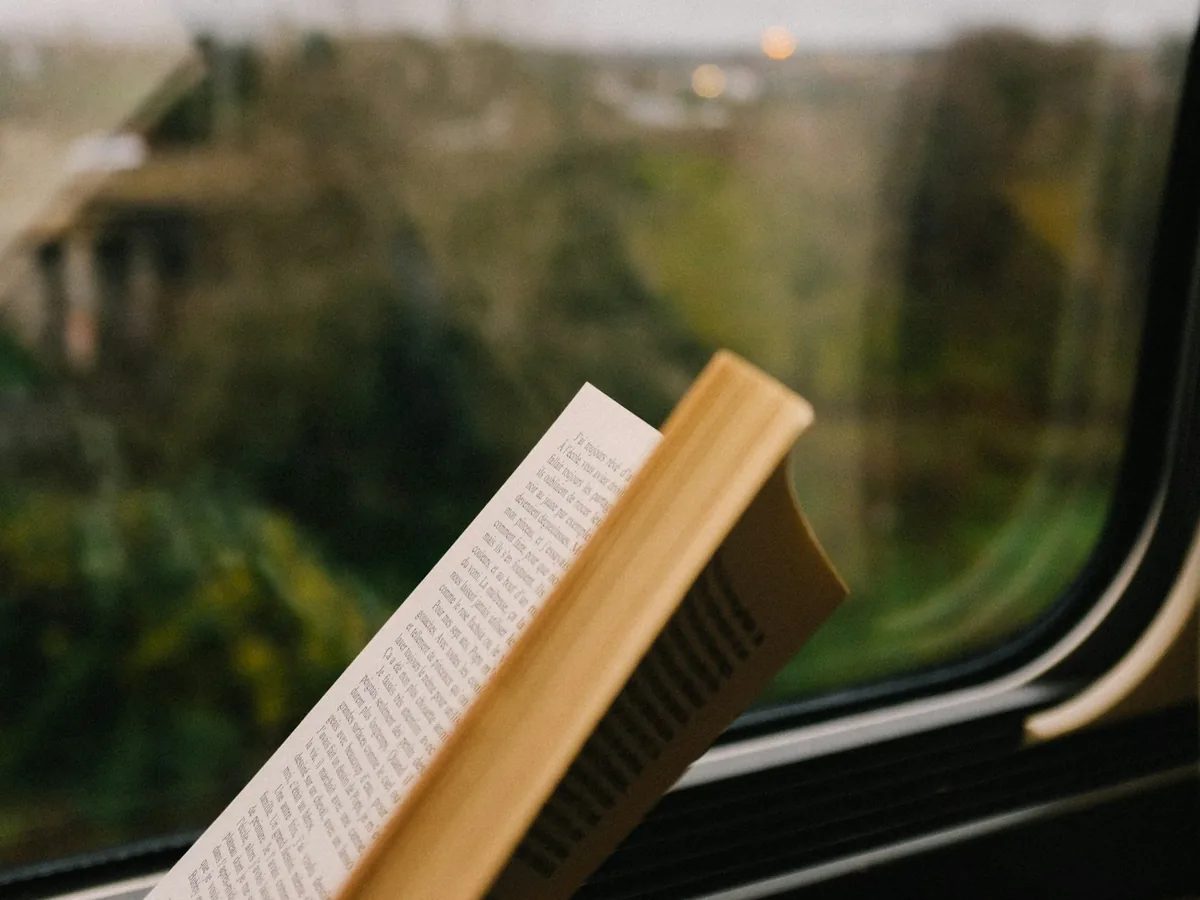 Un libro abierto sujeto junto a la ventanilla de un tren con colinas verdes pasando en el exterior
