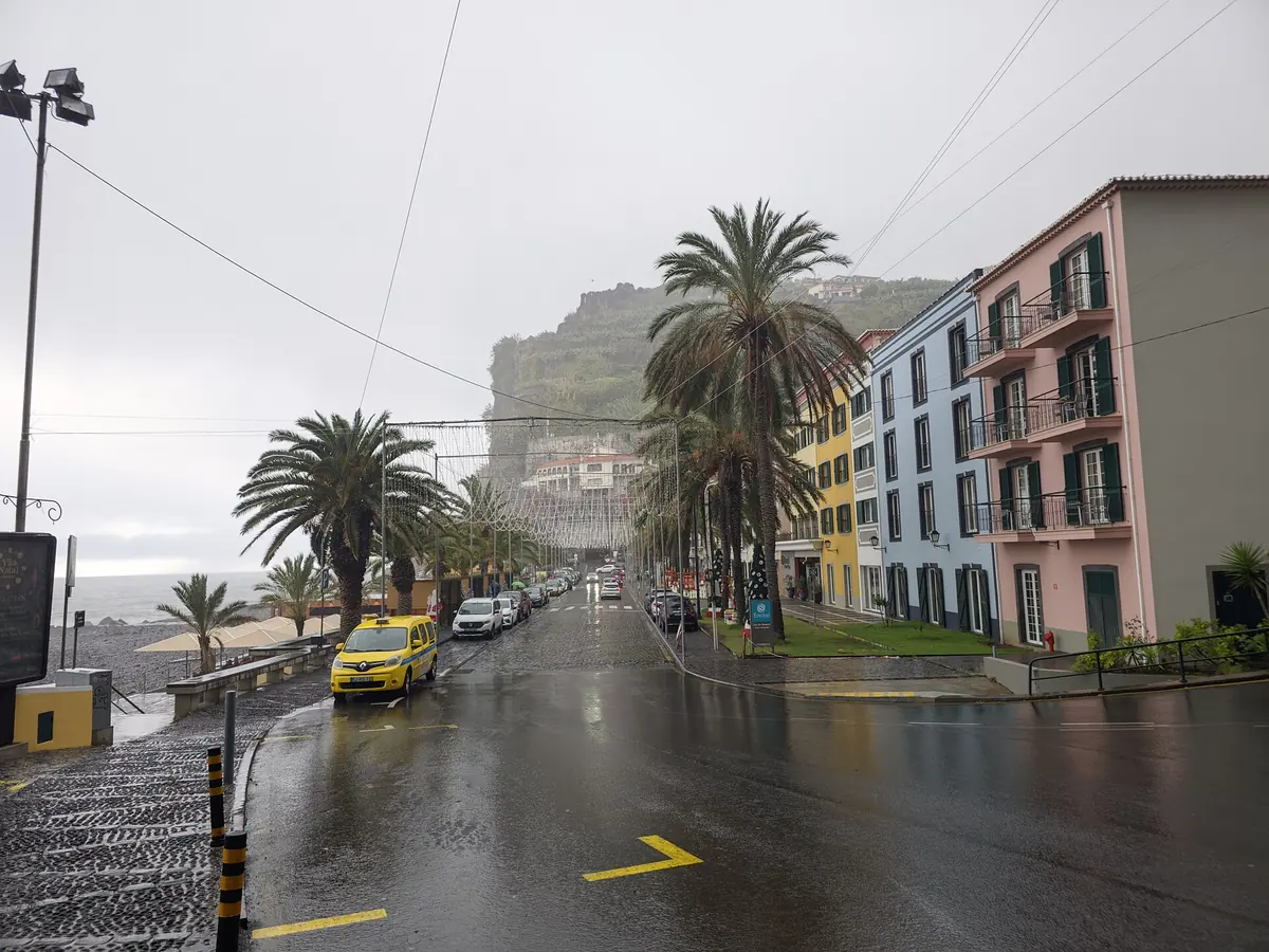 Una calle lluviosa junto al paseo marítimo de Ponta do Sol, en Madeira, con palmeras, casas de colores y un acantilado detrás