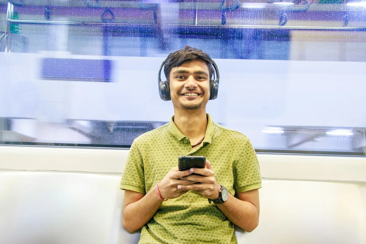 A smiling young man wearing black over-ear headphones looking at his phone on public transport