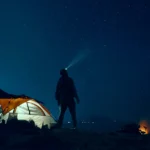 Ein Mann im Lichtstrahl seiner Stirnlampe neben einem leuchtend orangen Zelt unter einem Sternenhimmel
