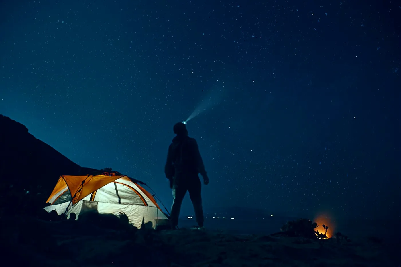 A man in the beam of his headlamp next to a glowing orange tent under a starry sky