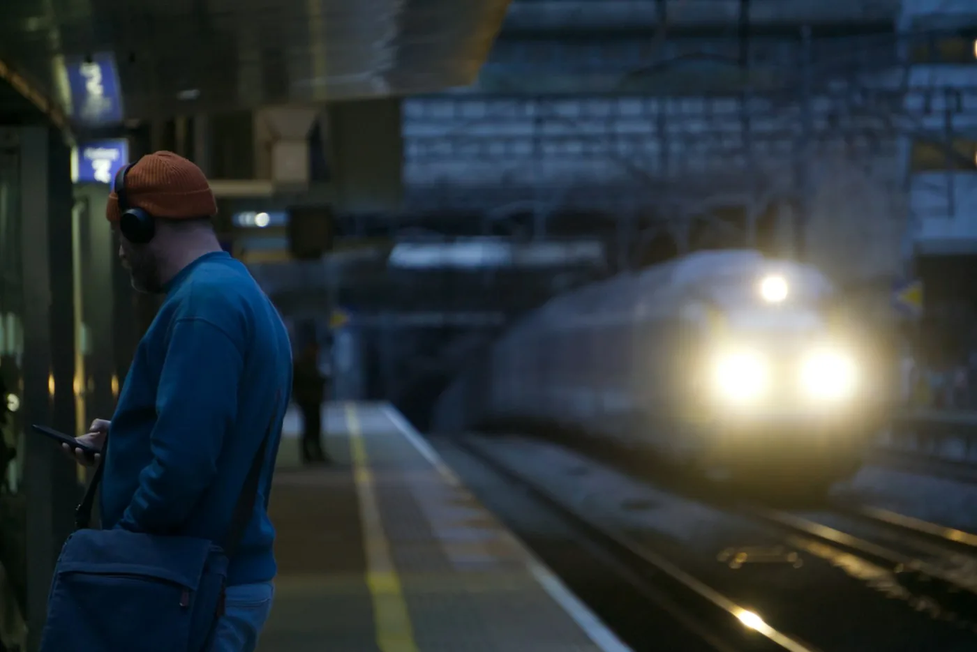 Man in headphones and a blue jacket waiting on a train platform at dusk as a train approaches
