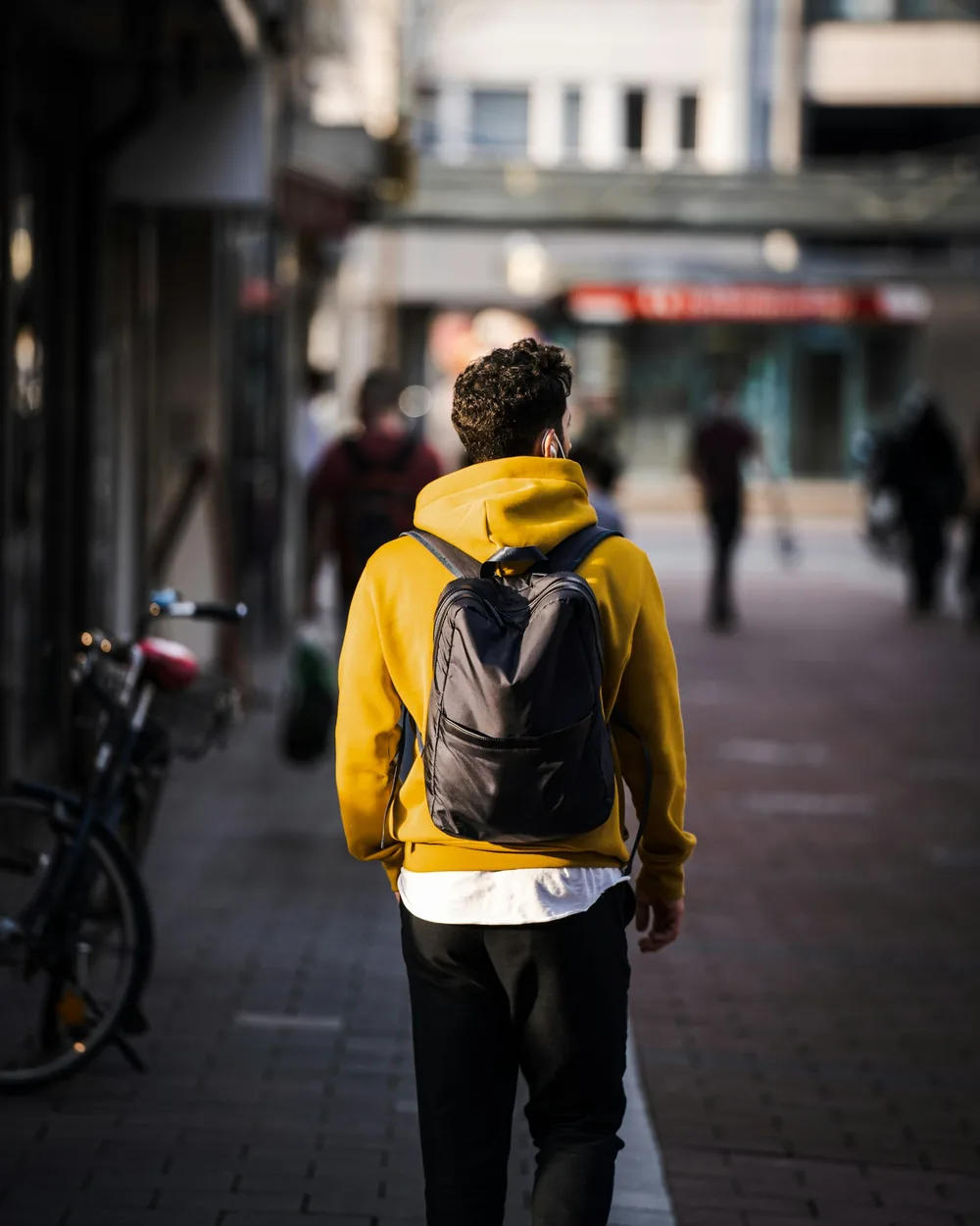 Un hombre con sudadera amarilla caminando por una acera de la ciudad con una mochila de día negra
