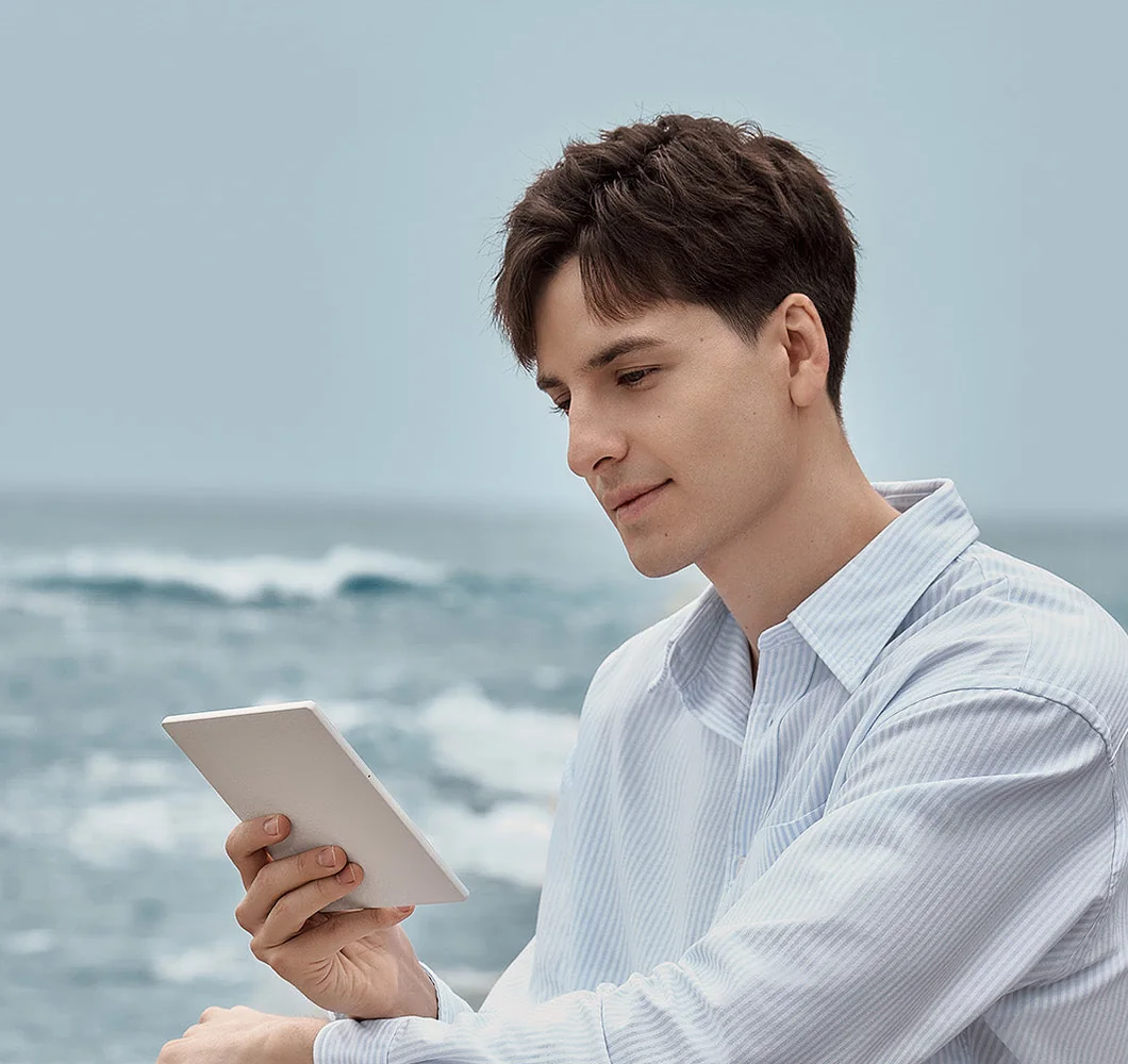 A man in a pale shirt reading a BOOX e-ink tablet on a rocky shore with waves behind him