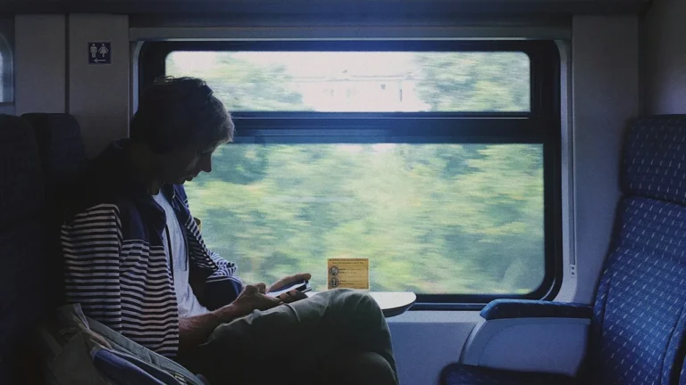 A man in a striped jacket sitting by a train window with green fields blurring past outside