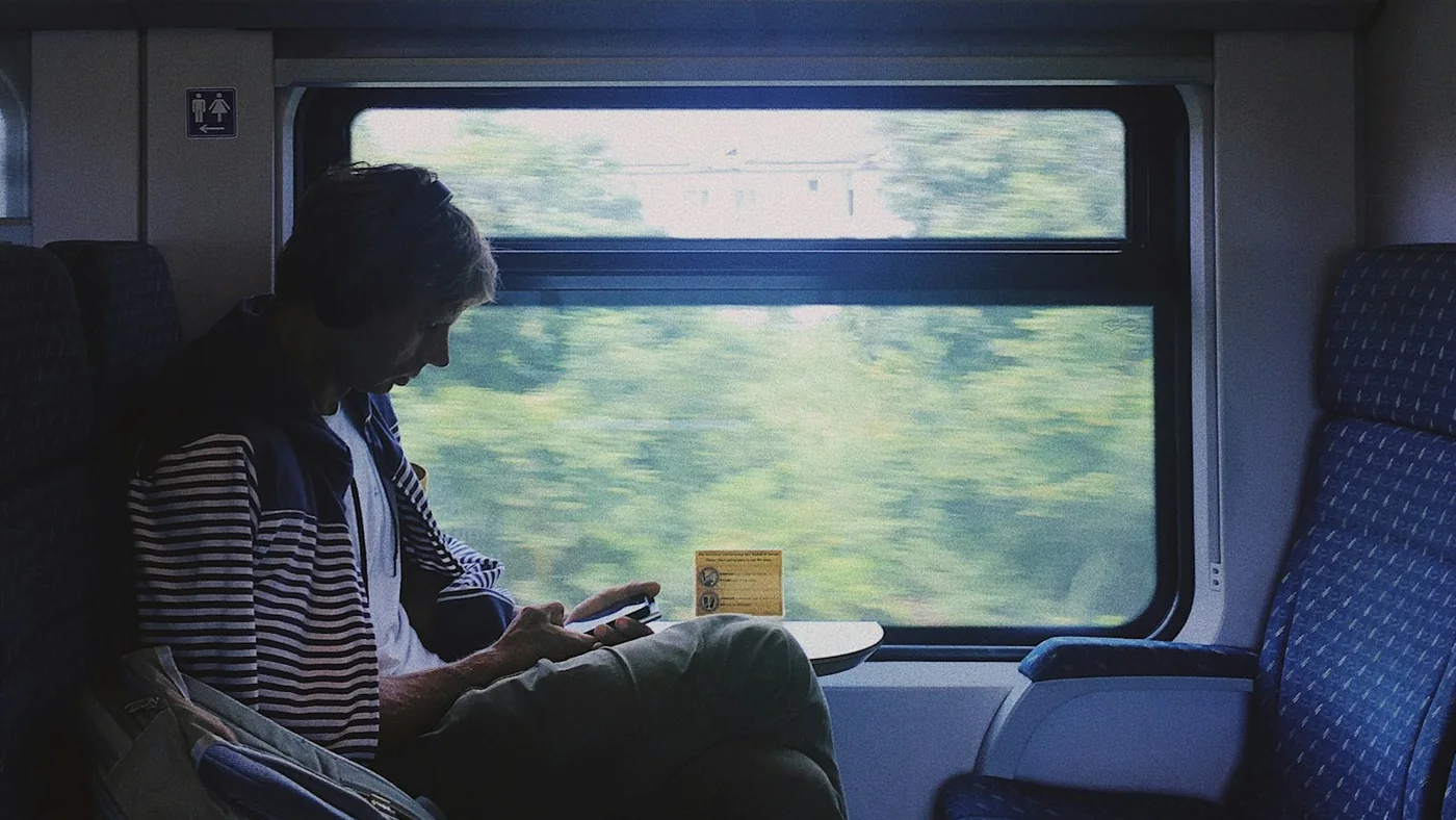 A man in a striped jacket sitting by a train window with green fields blurring past outside