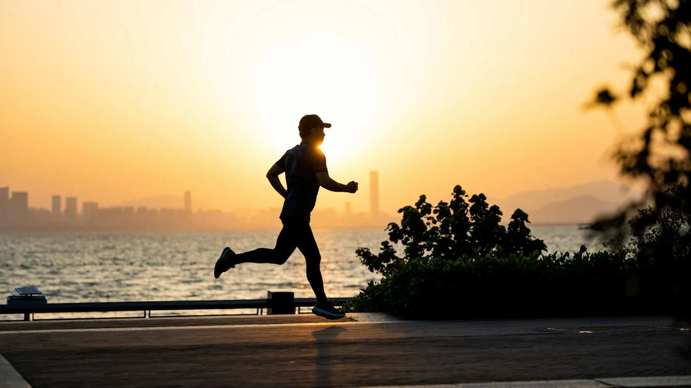 Silhouette of a man running along a seafront path at sunset with a city skyline across the water