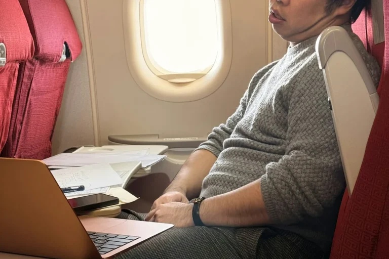 A man asleep in an airplane window seat with a laptop on the tray table