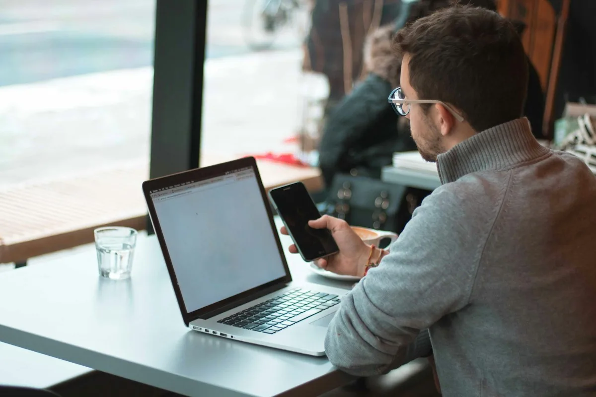 Man at a cafe window table with a laptop and a smartphone, using AI tools while working remotely